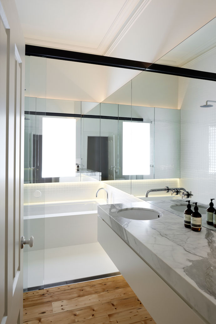 View to the bathroom at Heritage part of Hawthorn House. New white bathroom joinery and finishes delineates the new insertions to the old. Full room length mirror cabinet wall expands the narrow space. 